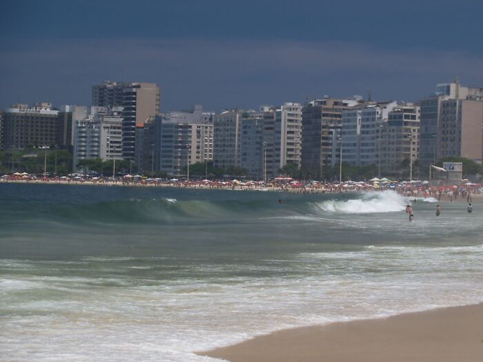 Brazilian beach scene with waves, city skyline, and beachgoers under cloudy skies.