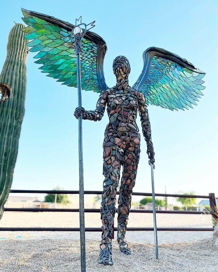 Winged angel sculpture with metallic texture holding a staff, showcasing interesting creepy handmade crafts against a clear sky.