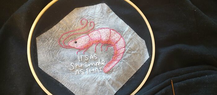 Embroidery hoop with a beautifully stitched pink shrimp and white text showcasing incredible embroidery skills on fabric.