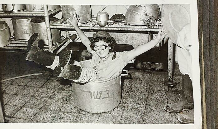 Child wearing boots and glasses playfully sitting inside a large pot in a kitchen, a candid glimpse into the past.