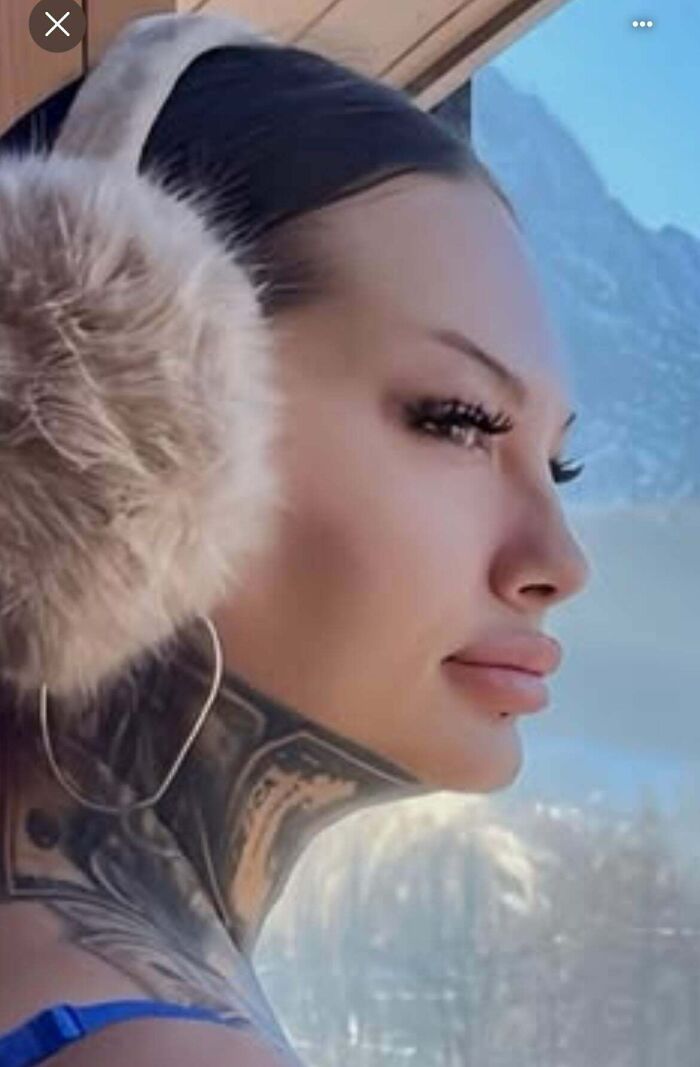 Person with airbrushed makeup, large furry earmuffs, and visible tattoos, gazing out at snowy mountain scenery.