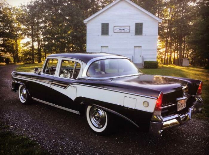 Vintage car parked outside a rustic white building, surrounded by trees at sunset.