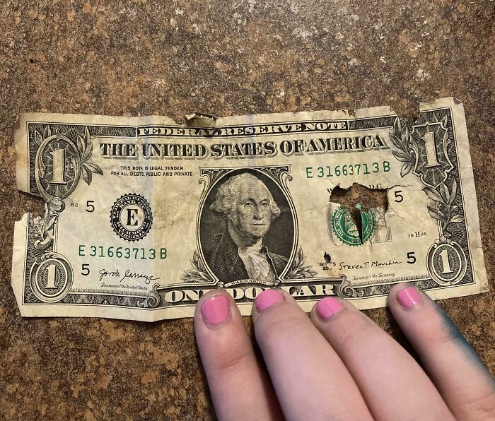 A worn dollar bill with a nail hole, resting on a surface, representing mildly infuriating server tips.