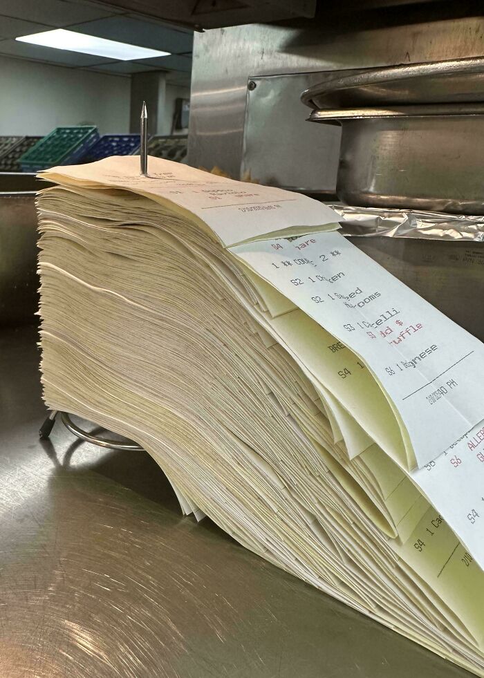 A stack of food service order tickets pierced by a spindle in a restaurant kitchen setting.