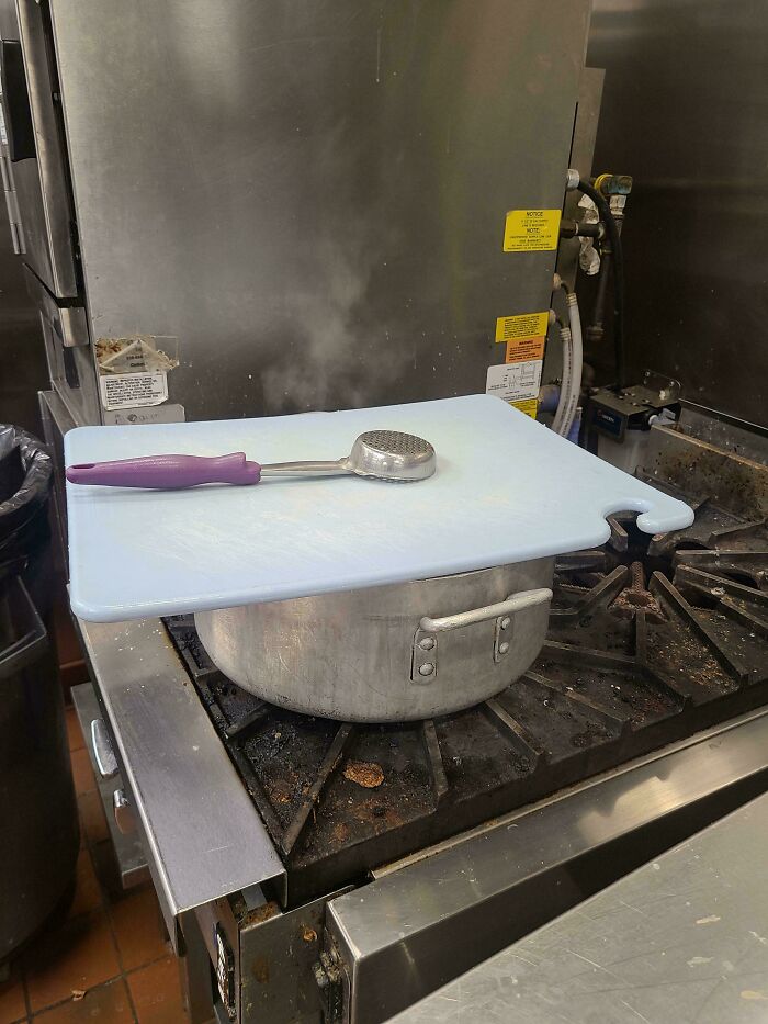 Improvised lid setup in food service kitchen, with pot, cutting board, and meat tenderizer on stove.
