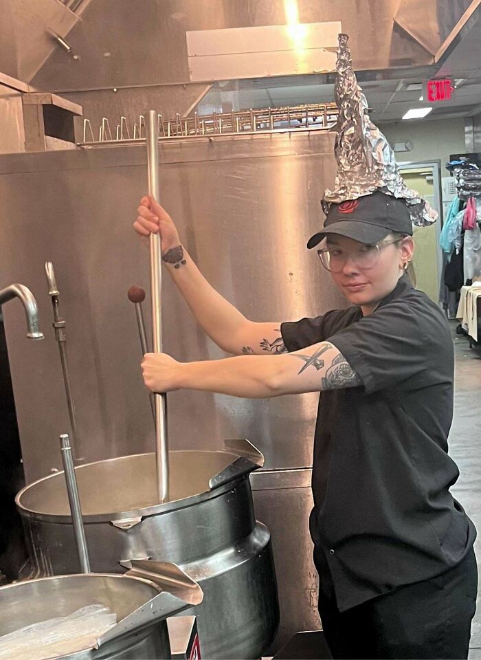 Person in a food service kitchen stirring a large pot, wearing a foil hat and glasses.