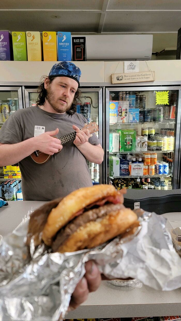 Food service worker playing ukulele behind counter with sandwich in foreground.