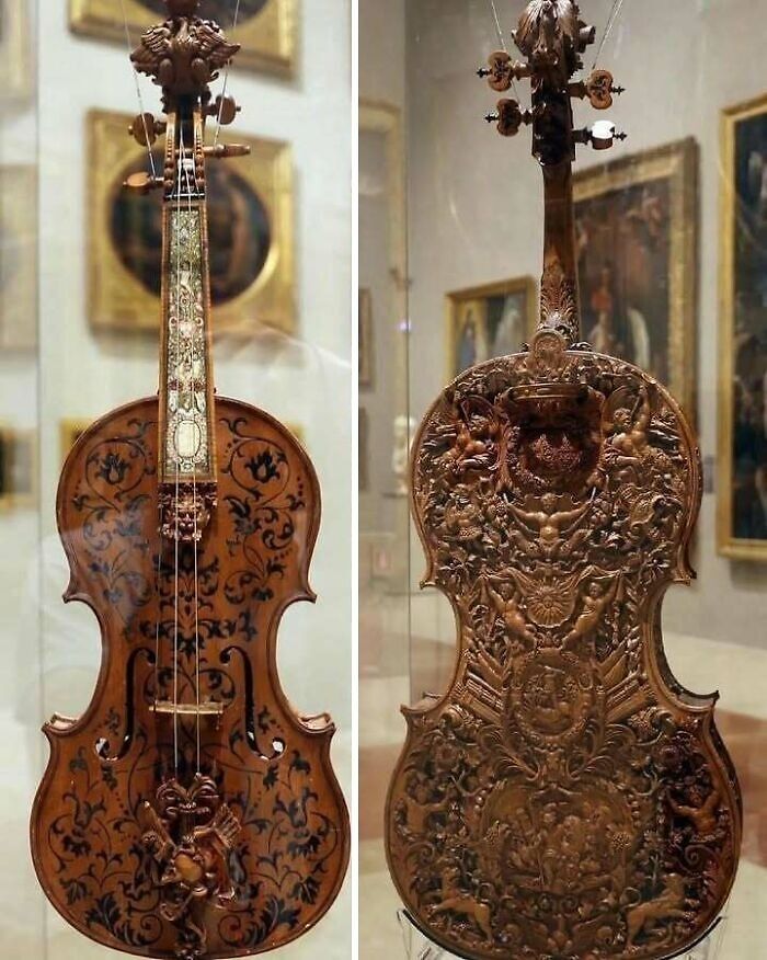 Intricately carved historical violin displayed in a museum, showcasing detailed artwork on both front and back surfaces.