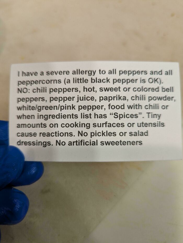 Allergy warning card for food service industry; lists pepper and spice allergies, held by a person in blue gloves.