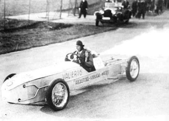 Inventor driving early race car, showcasing a creation that led to their own undoing.