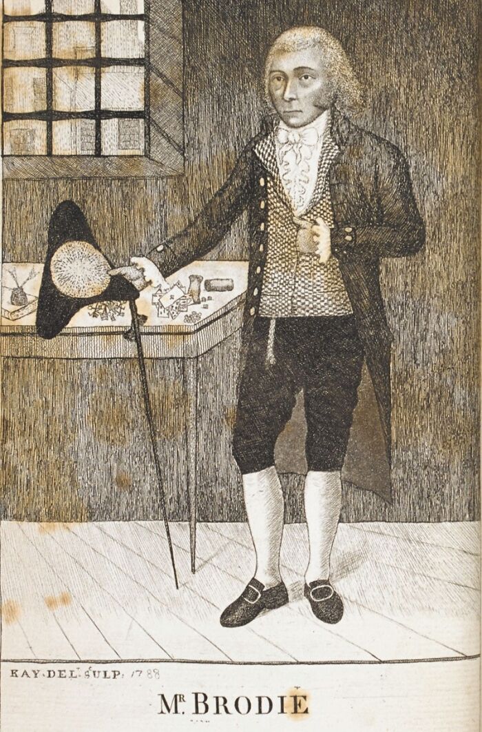 Inventor in 18th-century attire standing indoors, holding a hat and cane next to a table.