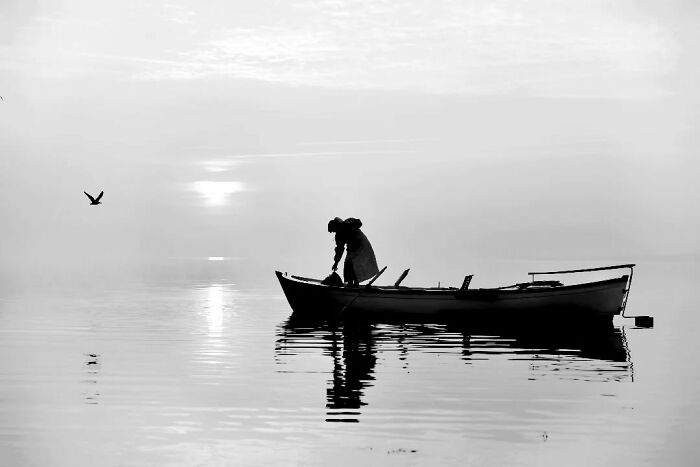 A serene black-and-white photograph captures a fisherman in rural Turkey on a boat at sunrise.