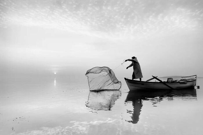 Fisherman casts net from a boat on a serene lake in rural Turkey, captured in stunning black-and-white.
