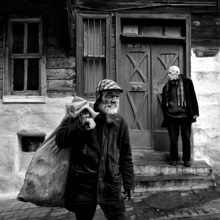 Elderly man carries sack in rural Turkey, captured in stunning black-and-white, with another man nearby in front of a wooden house.