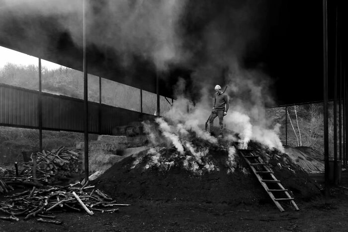 Black-and-white photo of a person standing on a smoking mound in rural Turkey, surrounded by wooden beams and logs.