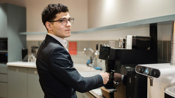 Man in an office kitchen using a coffee machine, wearing glasses and a suit; illustrating rules made due to one individual.
