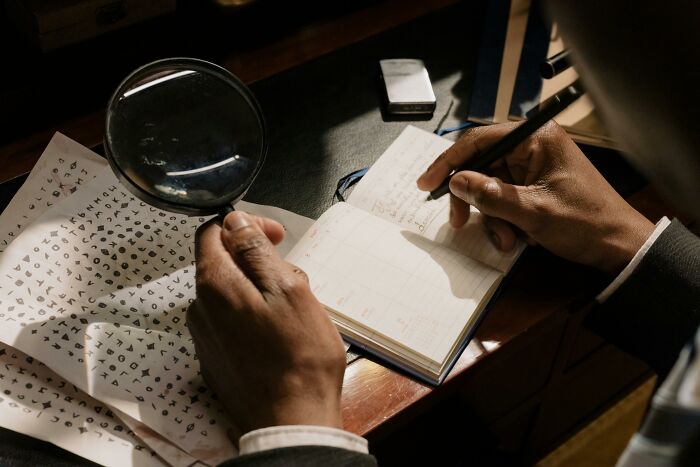 Person examining encrypted papers with a magnifying glass, jotting notes. Rumors explored through written code and analysis.