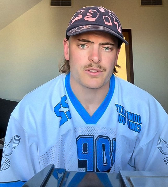 Podcaster in casual attire and cap, sitting indoors, during an emotional moment.