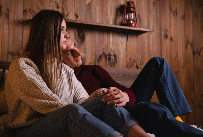 A couple sitting on a couch in a cozy cabin, contemplating uncovering background secrets.