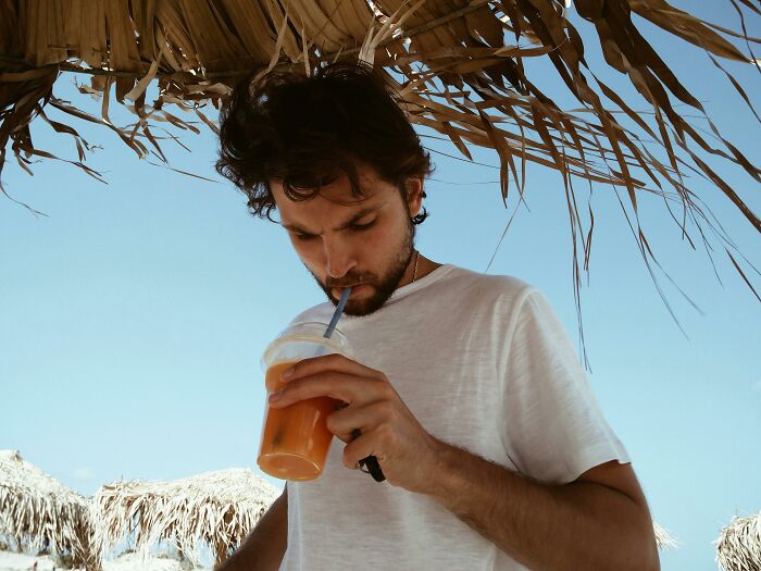 Young man drinking juice under a thatched umbrella, reflecting on notions of what real men don’t do in society.