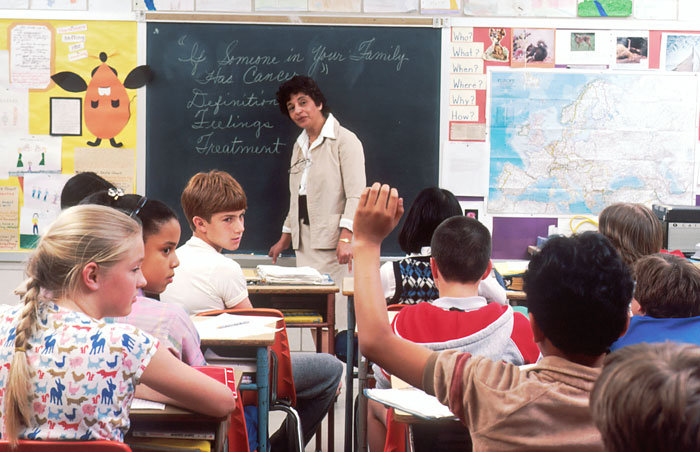 Estudiantes en clase mientras la profesora explica, un alumno levanta la mano para decir algo gracioso.