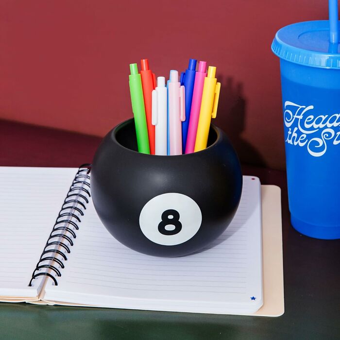 Bando pen holder shaped like a black eight-ball, with colorful pens on an open notebook beside a blue cup.
