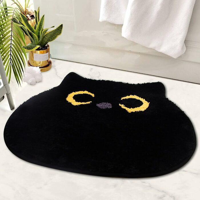 Black cat-shaped bathroom mat with yellow eyes on a white tiled floor near plants and a towel hanging.