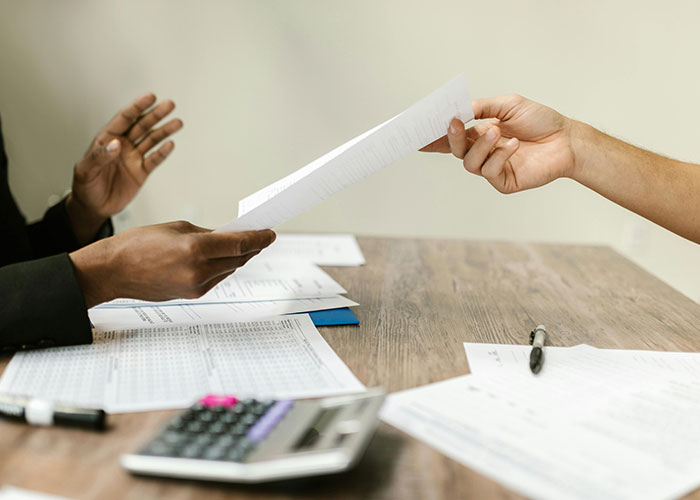Two people exchanging documents across a table with charts and a calculator, illustrating workplace dynamics and potential revenge.