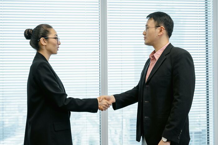 Two professionals in business attire shaking hands, discussing rumors in an office setting.