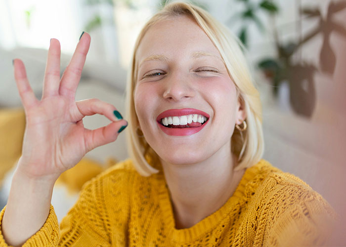A woman winking and smiling, wearing a yellow sweater, while making an okay gesture, capturing a whimsical moment.