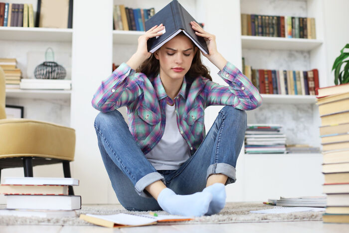 Person sitting with a book on their head, surrounded by books, depicting American norms in a library setting.