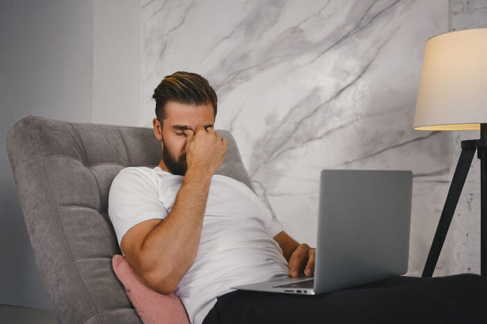 Man in white shirt sitting on a chair, pinching nose in frustration while looking at a laptop, dumbfounded expression.