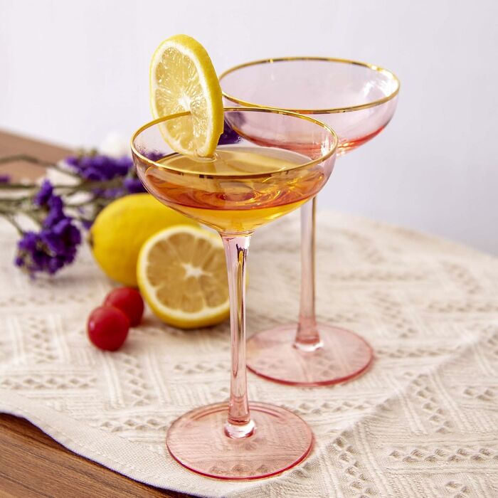 Two elegant pink-rimmed cocktail glasses with lemon garnish on a lace tablecloth, showcasing expensive taste.