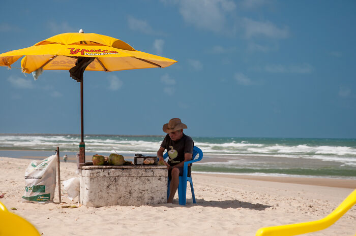 Man sitting under a yellow umbrella on a Brazilian beach, with coconuts on a table.