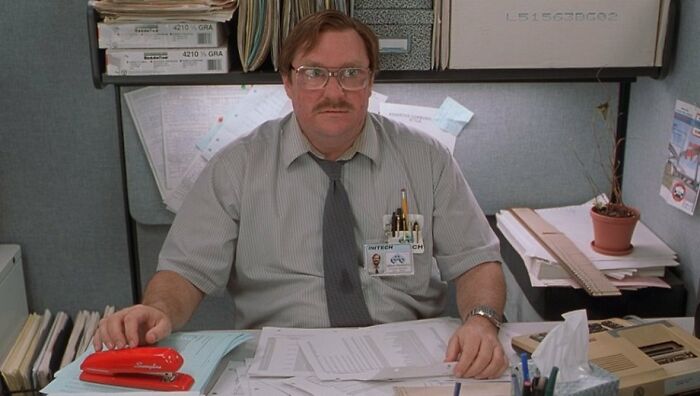 Office worker with glasses at cluttered desk featuring famous movie props like a red stapler.