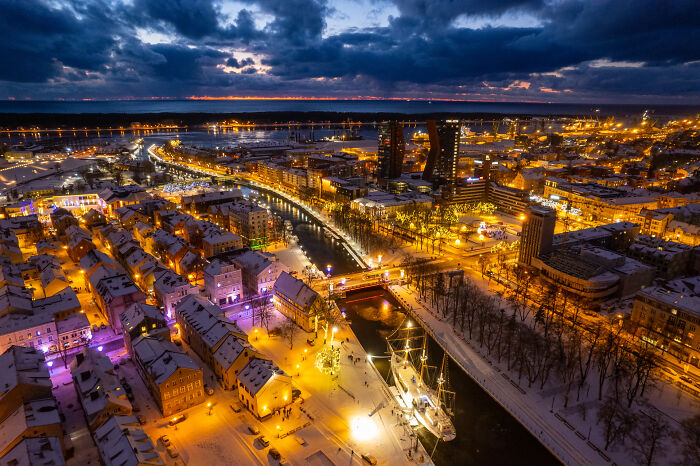 Lithuania bird’s-eye view at dusk, showing illuminated streets, buildings, and a river with a ship, under a dramatic sky.