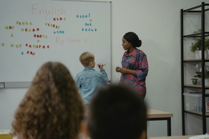 Child writing on a whiteboard in English class while a teacher supervises, illustrating professions people often underappreciate.