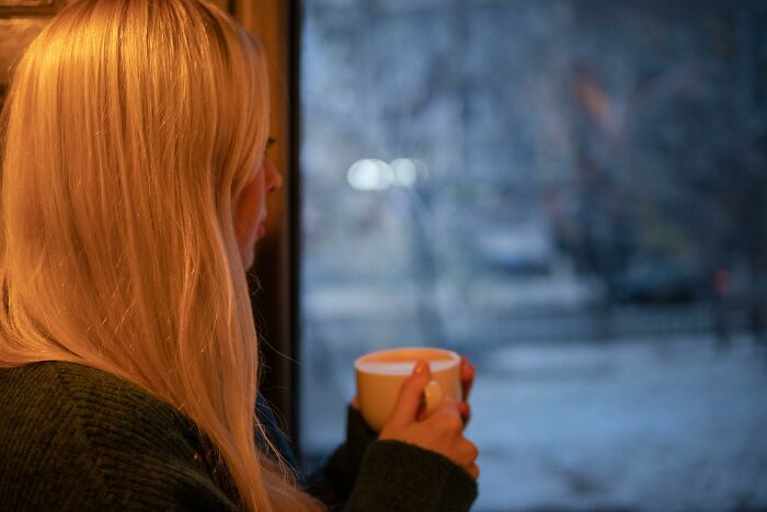 Woman with long blonde hair enjoying a warm drink by the window, highlighting perks of not going to the office.