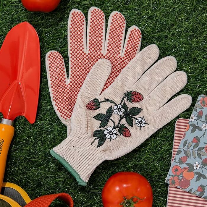 Gardening gloves with strawberry design from Bando on grass, surrounded by a trowel and tomato.