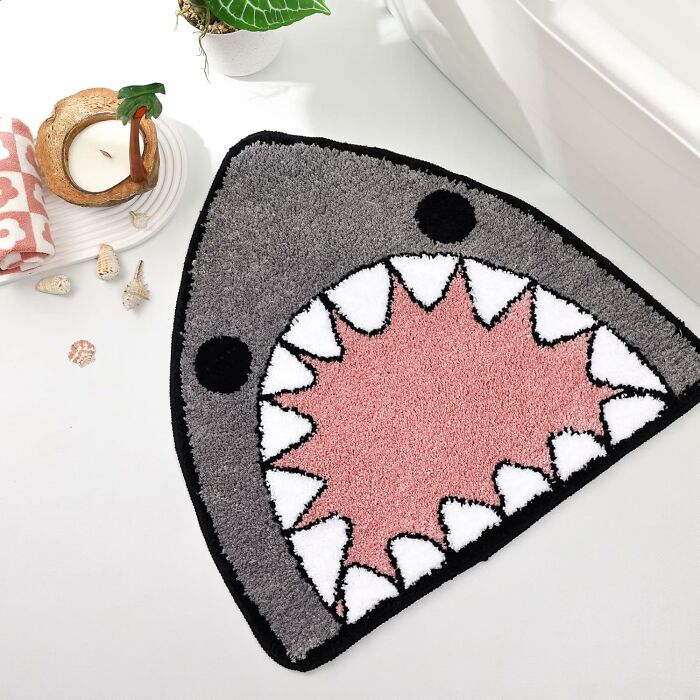 Shark-themed bathroom mat with gray, pink, and white colors placed near a white bathtub and decorative shells.