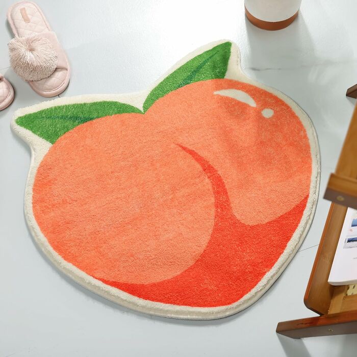 Peach-shaped bathroom mat with green leaves on a light floor next to pink slippers and a wooden chair.