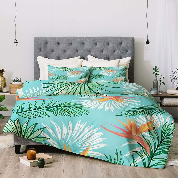 Tropical patterned bedding from Society 6 on a gray tufted bed with plants and candles nearby.