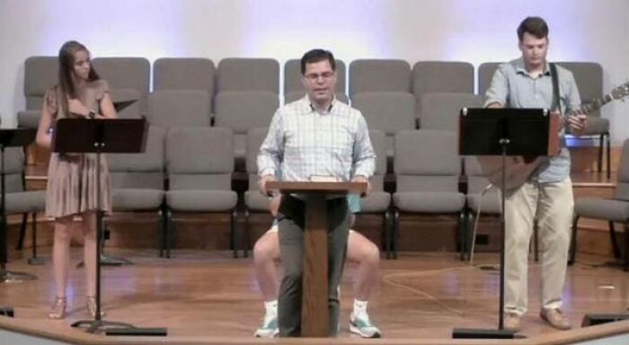 A man appears oddly seated at a podium with two legs in sneakers visible, accompanied by musicians on stage.