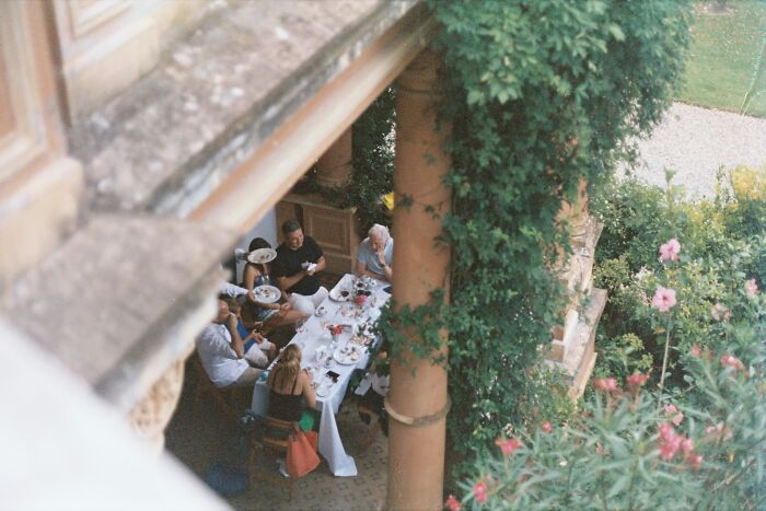 People gathered at an outdoor family event, sitting around a long table, surrounded by lush greenery and flowers.