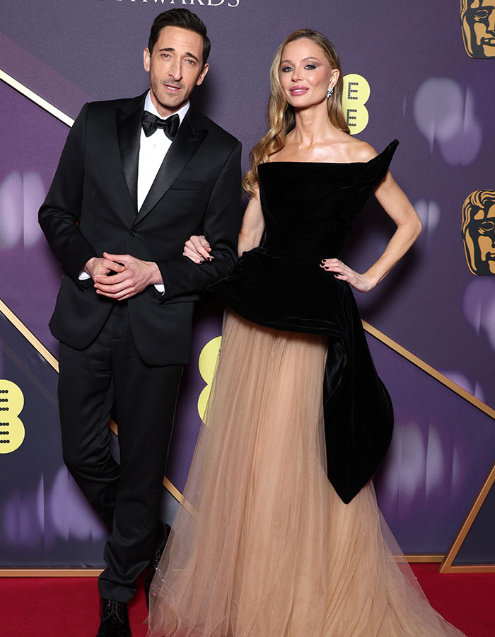 Man in a black tuxedo and woman in a black and beige gown on BAFTA red carpet, showcasing daring outfits.