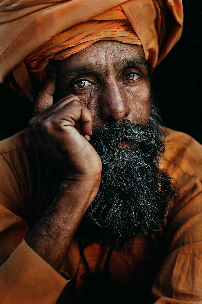 Expressive face of a man with intense eyes and a grey beard, wearing an orange turban, hand resting on his cheek.