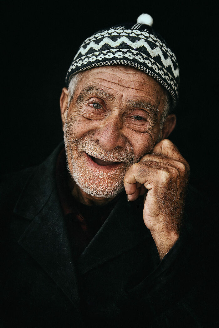 Expressive face of an elderly man in a patterned hat, smiling warmly against a dark background.