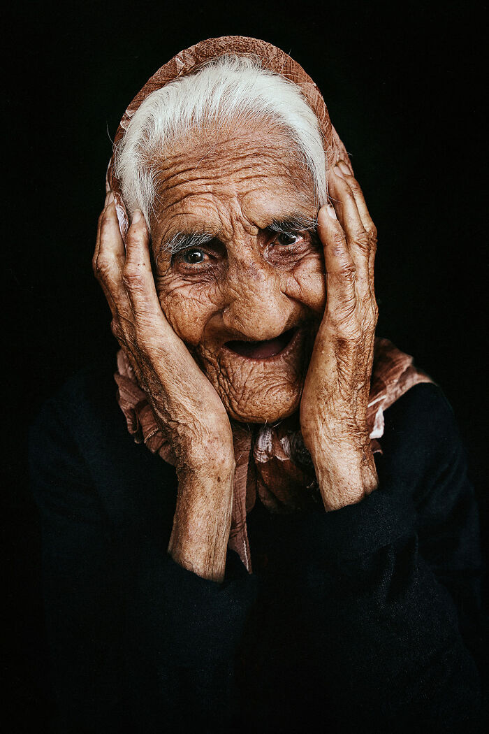 Photographer captures expressive face of elderly woman with hands on cheeks against dark background.