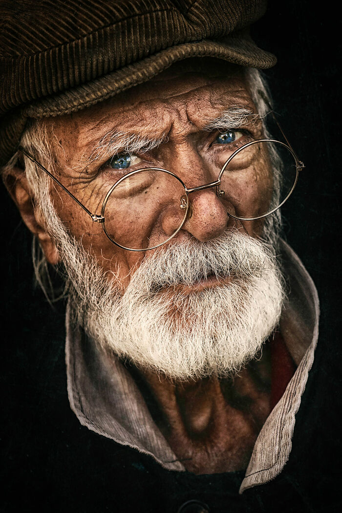 Expressive elderly man with glasses, wearing a hat and a white beard, captured by a photographer.