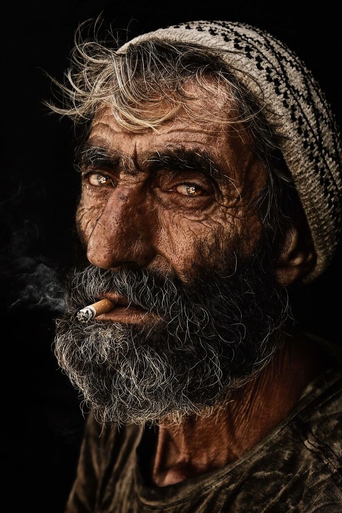 Expressive older man with a weathered face, wearing a knit cap, smoking a cigarette against a dark background.